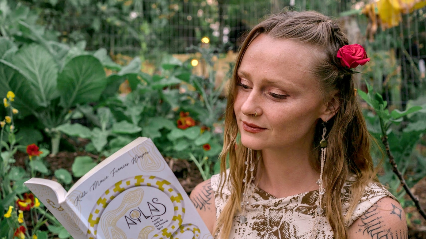 Poet Kelli Harper stands in a garden reading from her poetry book, Atlas. She has a pink flower in her blonde hair, which falls over her shoulders.