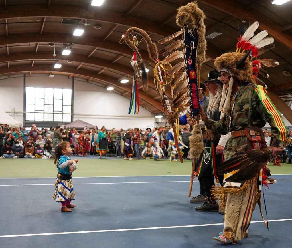 Photograph of a small child in regalia on the left facing two adult dancers in regalia on the right in an indoor tennis court arena.