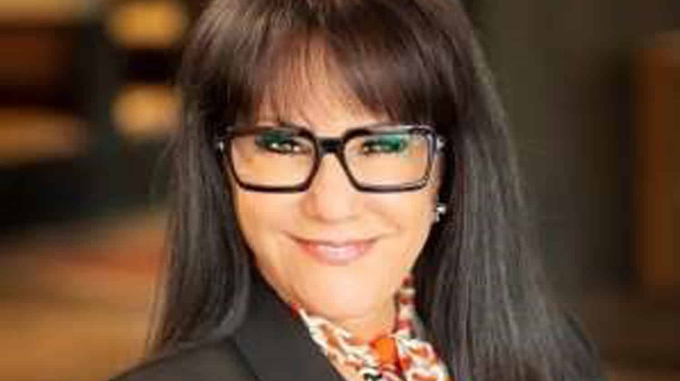 Headshot of Crissy Rumford in a black blazer and patterned blouse in warm colors. Her black glasses pop on her face, and her smile is energetic and bright.