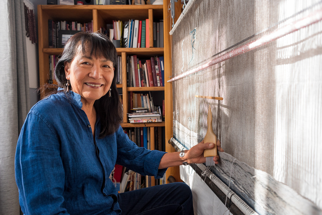 An artist wearing a dark denim shirt sits at a loom, turning towards the camera and smiling.