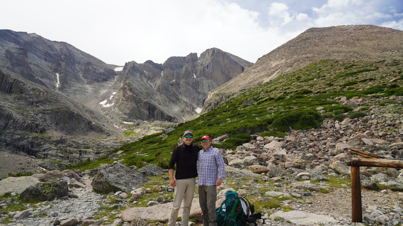 Father and son pose for a photo with enormous grey mountain peaks in the background.
