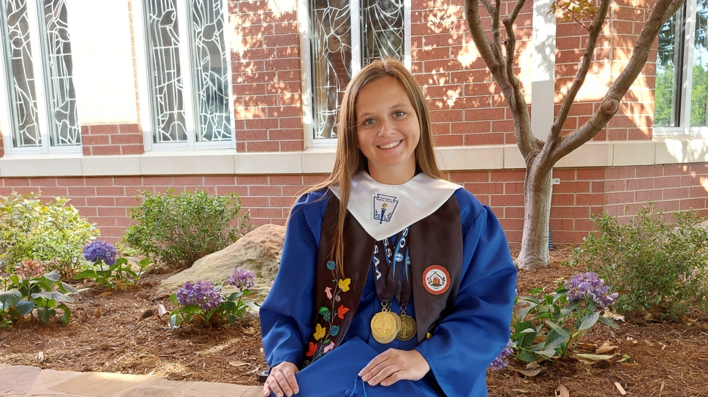 Tribal member Shelby O'Steen sits on a ledge in front of a brick wall. She wears a bright blue high school graduation gown and a CPN graduation stole, and holds her graduation cap in her hands.