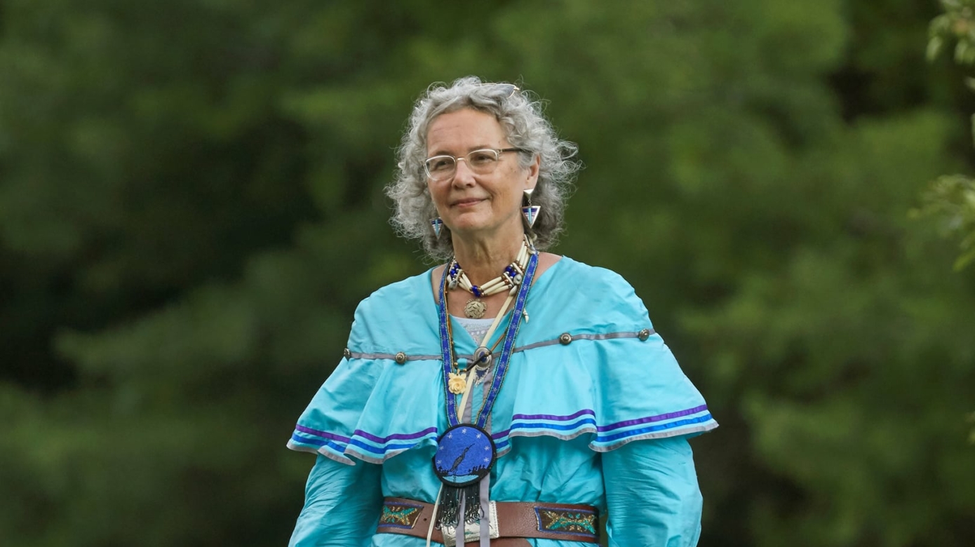 Full-body portrait of photographer and CPN Tribal member Sharon Hoogstraten in regalia of dark and teal blues with orange floral accents. She stands on a rock in front of some trees, and the light suggests a slight cloud cover.