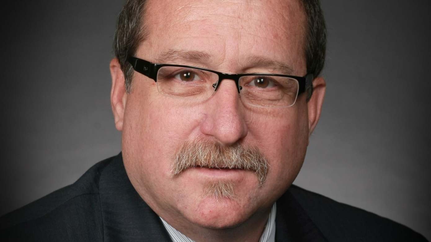 Headshot of Representative Mark McBride. He wears a suit and tie, and has dark-framed glasses on. A greying mustache matches a small goatee.
