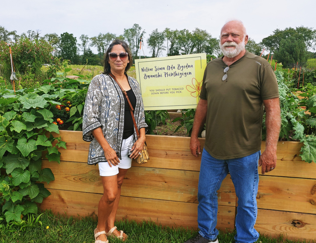 Two people stand on either side of a yellow sign in a large raised planter bed overflowing with plants and produce.