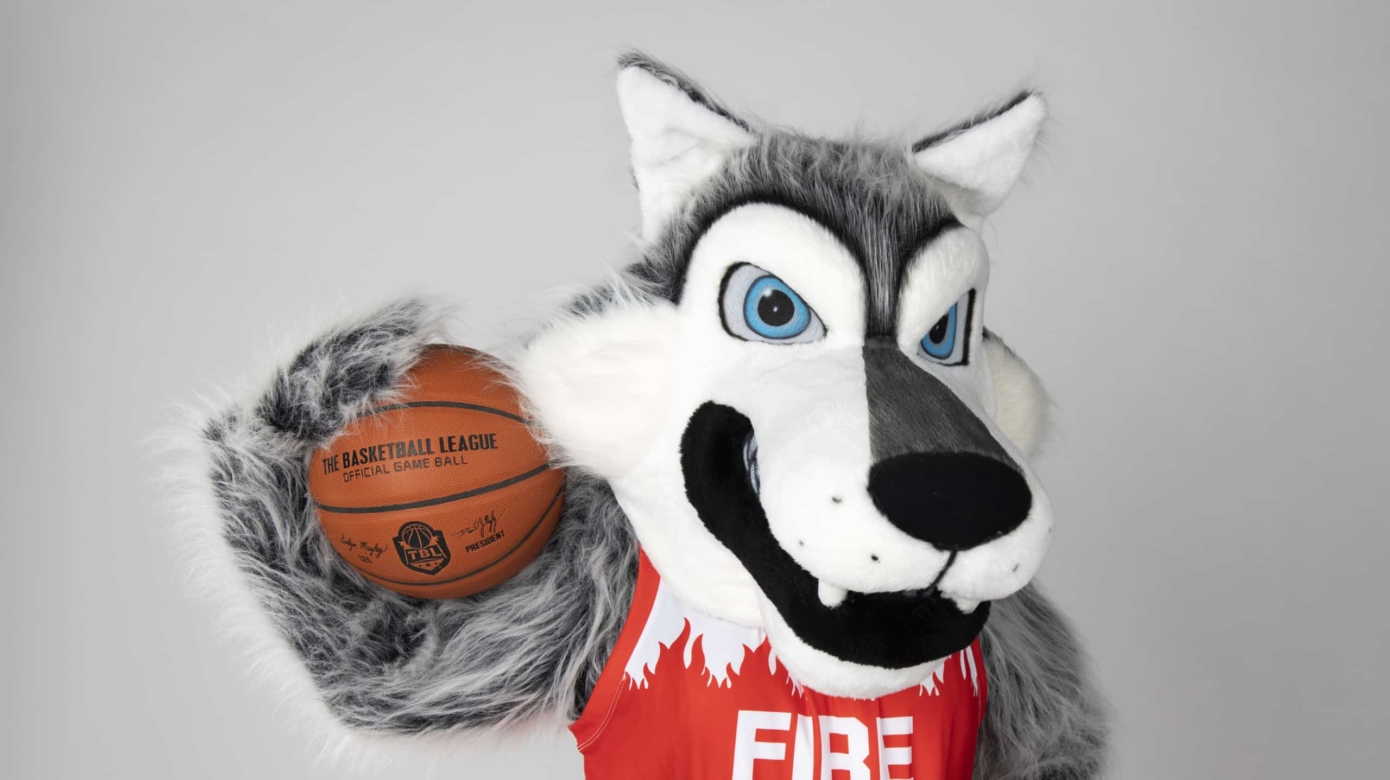 Potawatomi Fire wolf mascot, Mo, poses with a basketball on their shoulder. Mo wears a red Potawatomi Fire jersey.