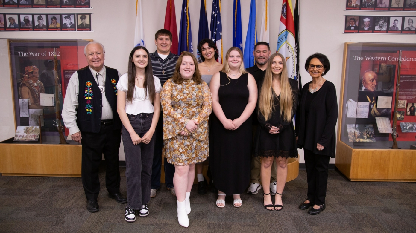 A group of students stands with Tribal Chairman John "Rocky" Barrett and Tribal Vice-Chairman Linda Capps in front of a flag display at the Nation's Cultural Heritage Center.