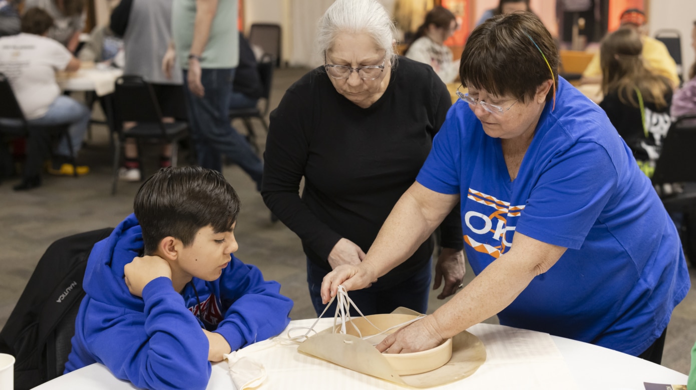 An instructor assists a student with making a hand drum. A Tribal elder watches.