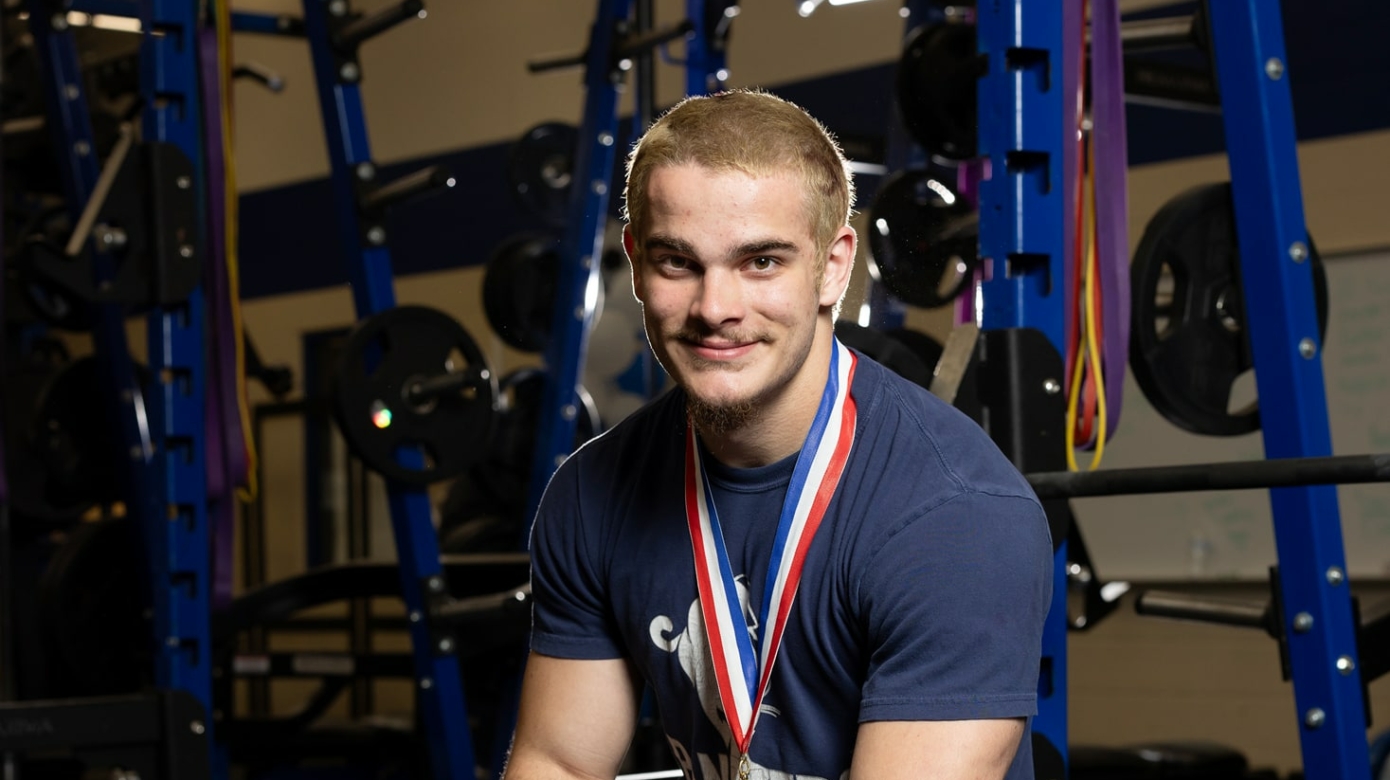 Kash Maynard sits with hands clasped in front of him on a weightlifting bench. He wears a gold medal around his neck.