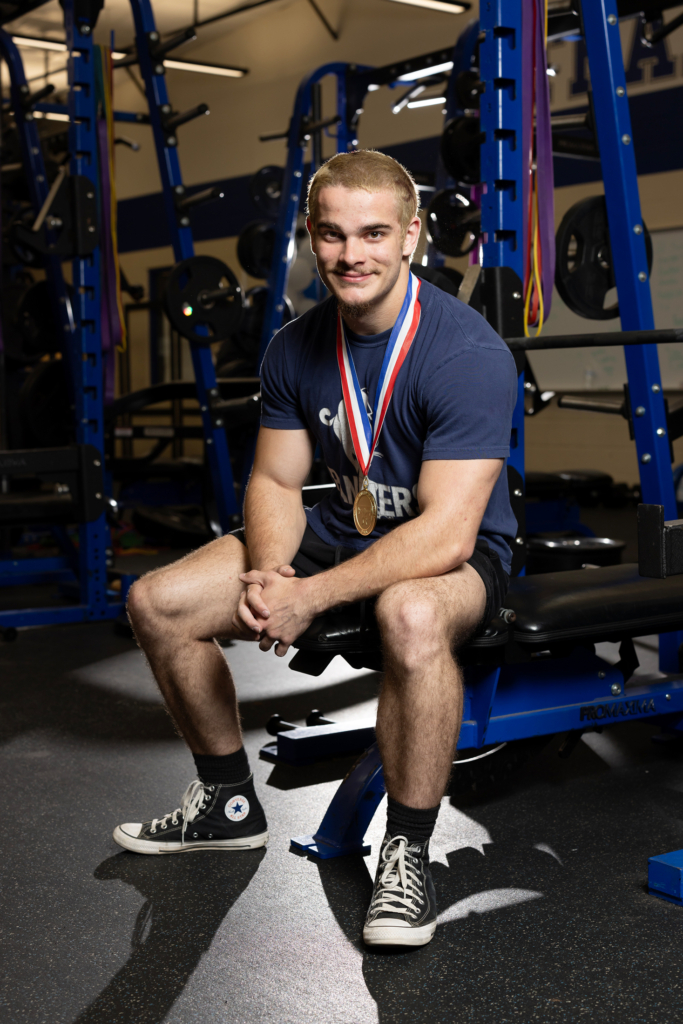 Kash Maynard sits with hands clasped in front of him on a weightlifting bench. He wears a gold medal around his neck.