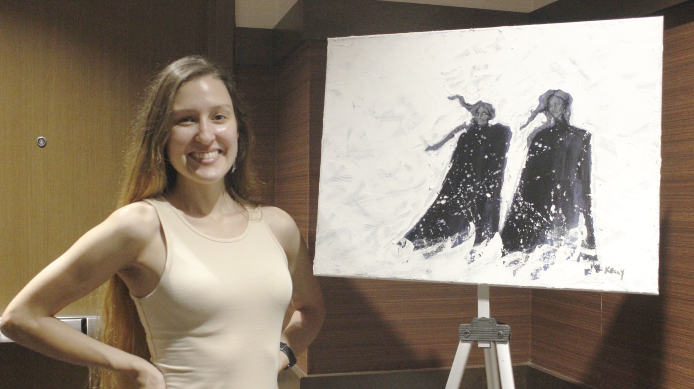 A young person with long brown hair poses next to a black and white artwork she created.