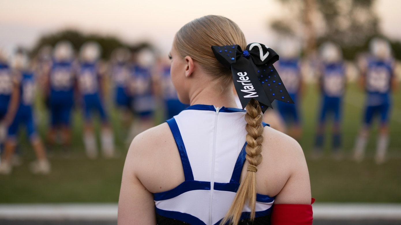 Marlee Affentranger stands with her back to the camera facing the football field. She wears a blue and white pom uniform and her blonde hair is braided in a neat ponytail with a bow bearing her name.