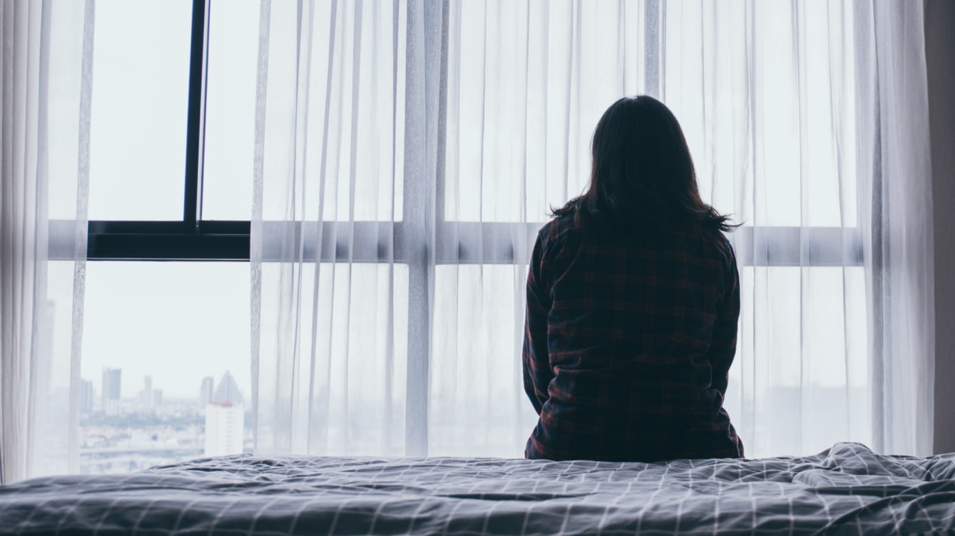 Silhouette of a person sitting on the edge of a bed in front of a window.
