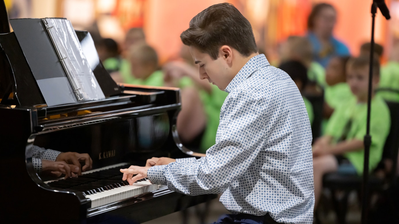 A young student in a blue patterned button down shirt plays a black piano.