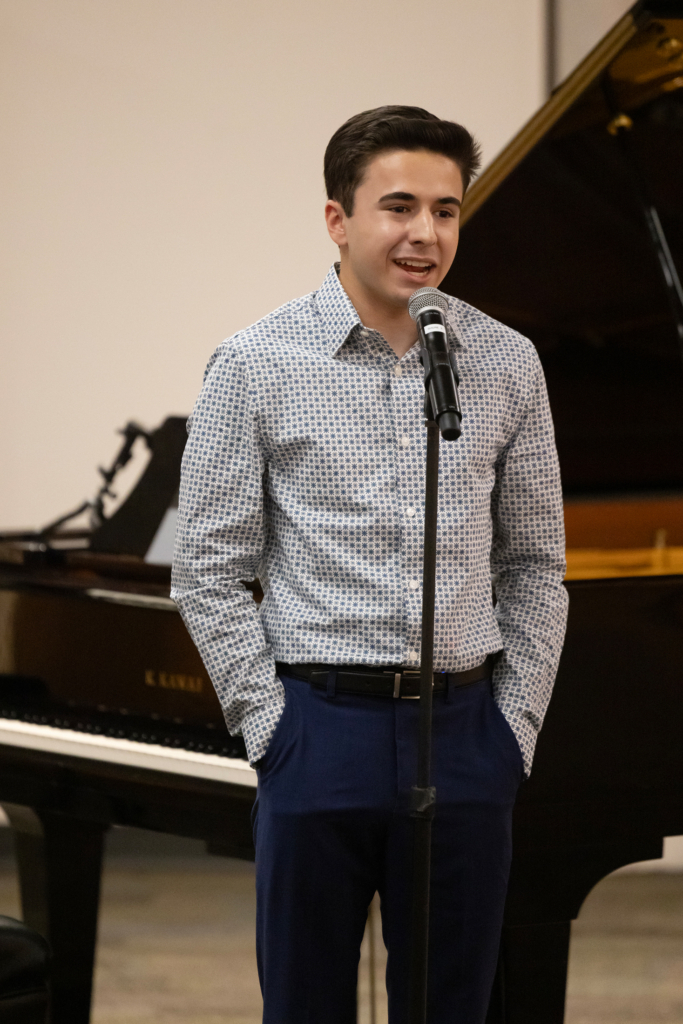 A young student in a blue patterned button down shirt and blue pants stands at a microphone to speak. 