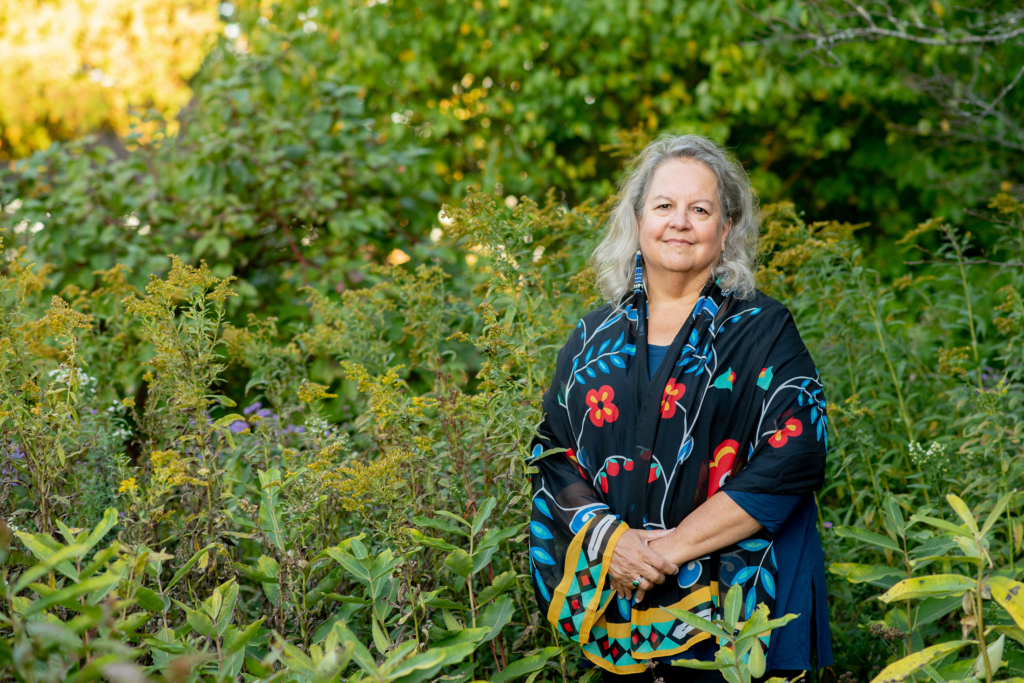 Dr. Robin Wall Kimmerer stands among vibrant green plants reaching to her shoulders. She wears a black shawl with brightly colored floral patterns on it.