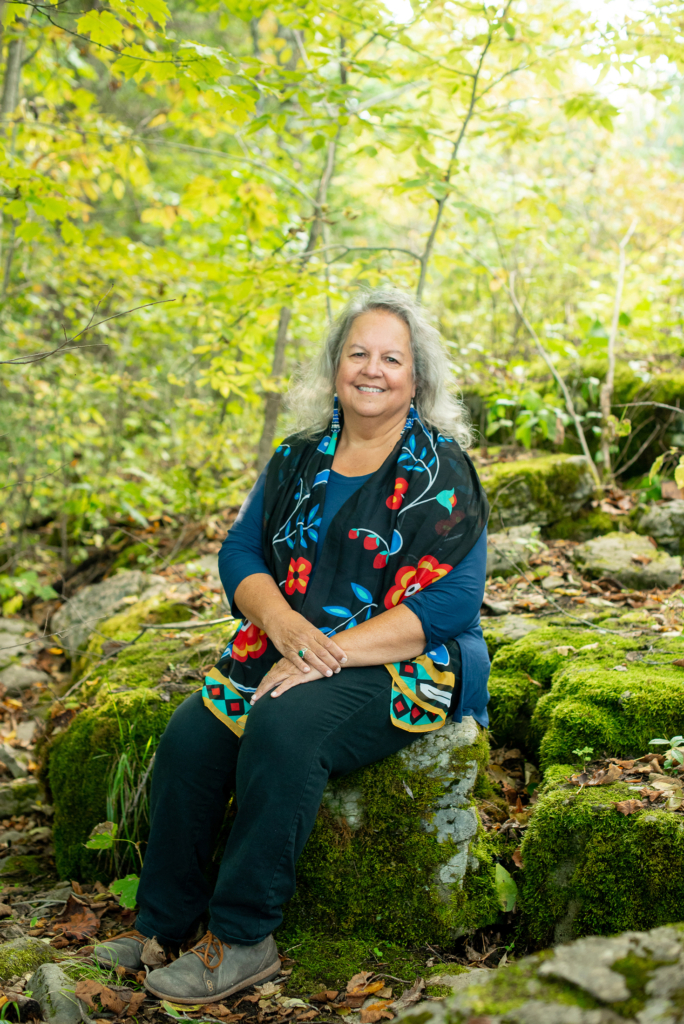 A woman with gray hair wearing blue jeans, a blue shirt, and a woodlands floral patterned scarf, sits on a mossy rock in a wooded area.